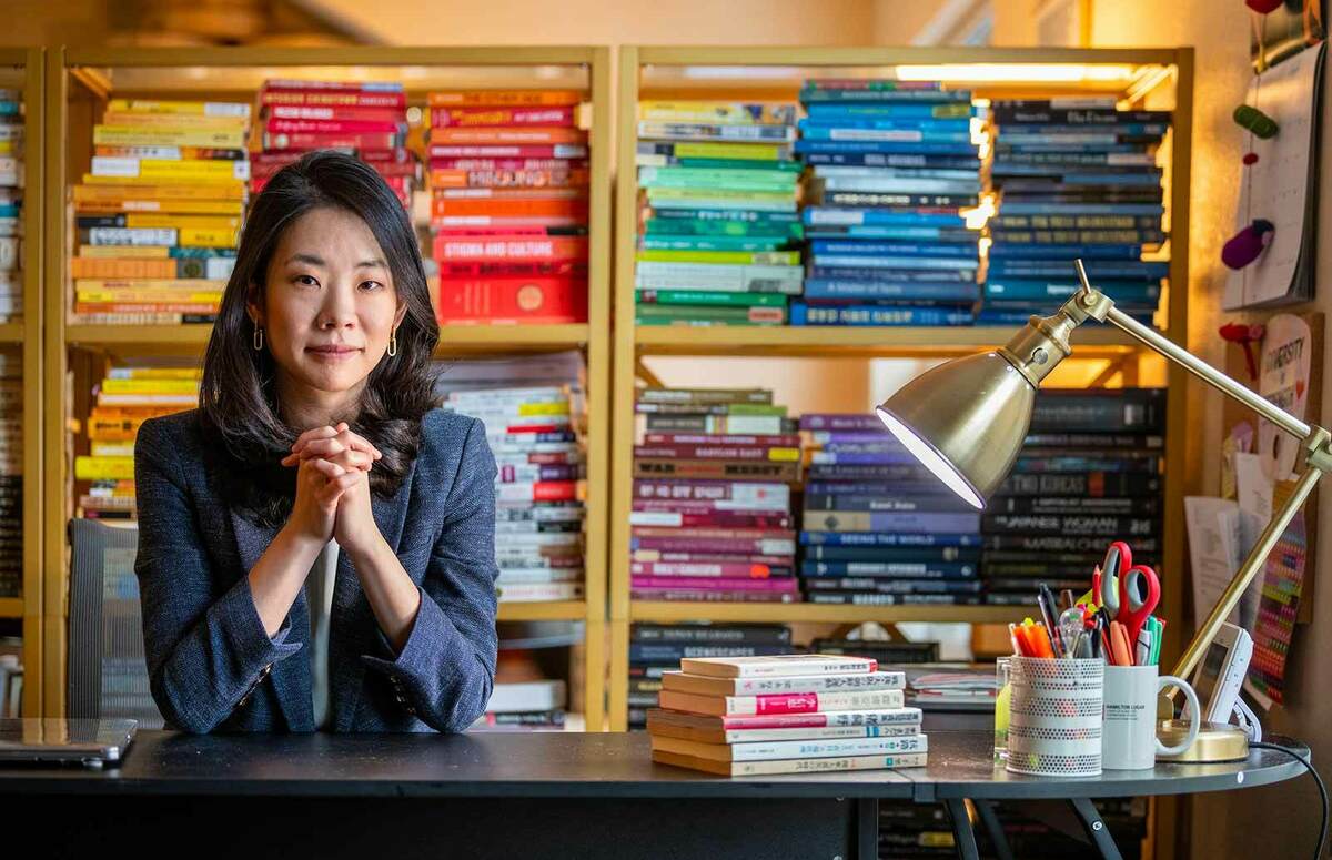 A woman poses for a photo behind her desk, both elbows on the table, and her hands create a relaxed fist. A bookshelf of color coordinated books behind her.