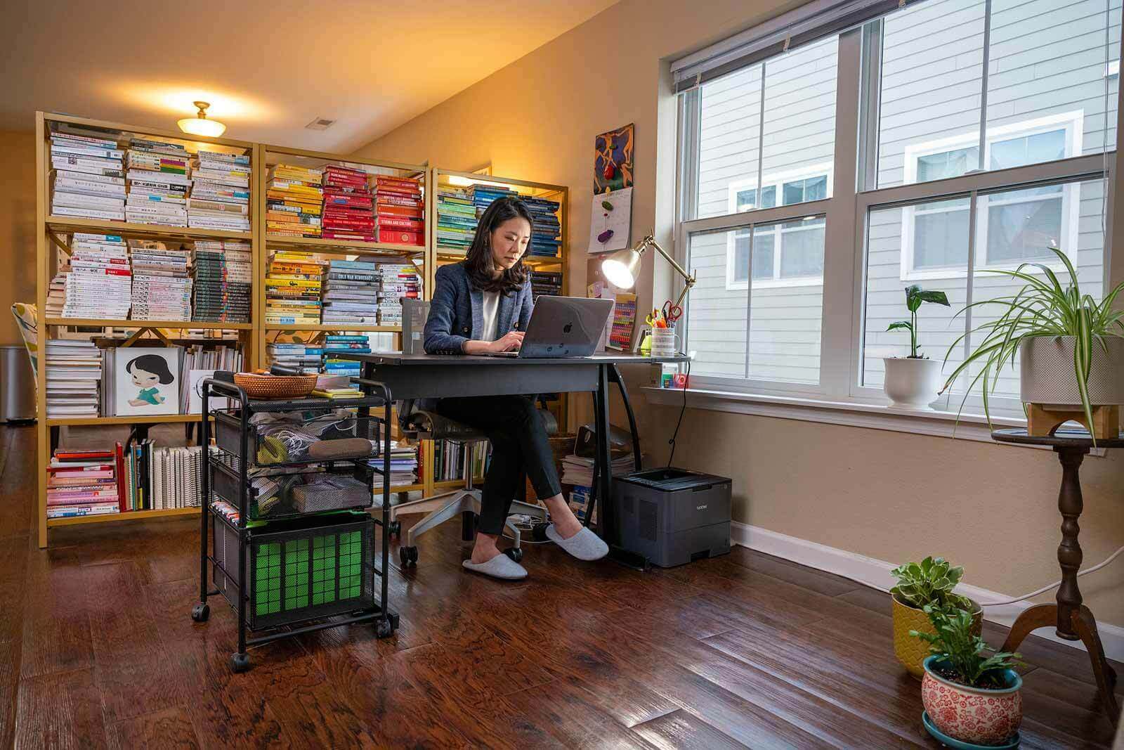 A woman sits at a desk, working on a laptop. A large book shelf is behind her full of color coordinated books.